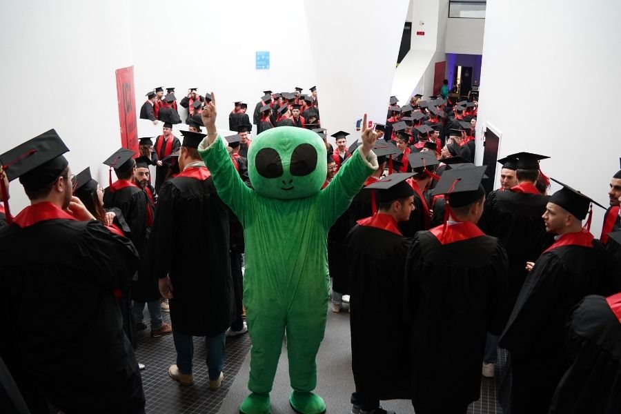 The UniLaSalle Amiens mascot “Alien” (a green extraterrestrial) raises his arms in the middle of 160 UniLaSalle Amiens graduates, dressed in American-style ceremonial attire (black gown, red sash, and black graduation cap).
