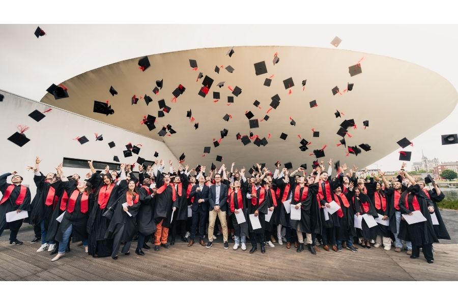 Group photo. The 160 graduates, dressed in formal attire (black gowns and red sashes), toss their hats in the air in front of the “flying saucer,” the iconic building shaped like an upside-down dome at UniLaSalle Amiens.