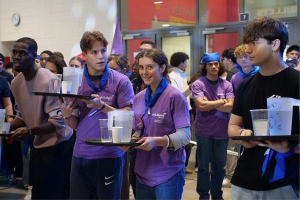2 étudiants lasalliens (un garçon et une fille), vêtus d'un t-shirt violet aux couleurs d'UniLaSalle Rouen, au départ de la course de garçons de café. Ils portent un plateau avec une carafe en plastique, et deux gobelets en plastique de taille différente. De part et d'autres,deux autres étudiants participants, issus d'autres établissements.
